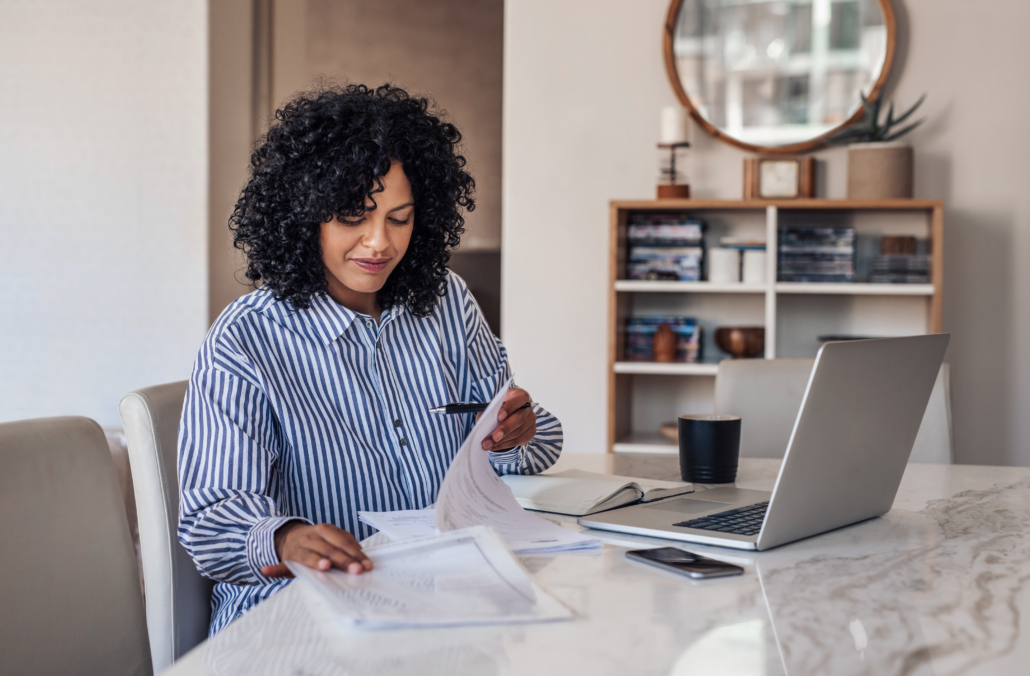 Smiling young female entrepreneur going through paperwork while working on a laptop at her dining room table at home